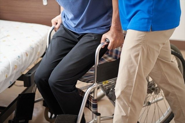 車椅子から立ち上がる高齢者を介助する介護士の男性.jpg